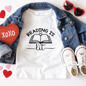 May include: White t-shirt with a black graphic that says "Reading is lit" with a book and heart design. The shirt is laid out on a white surface with a denim jacket, red heart-shaped sunglasses, and red and pink hearts.