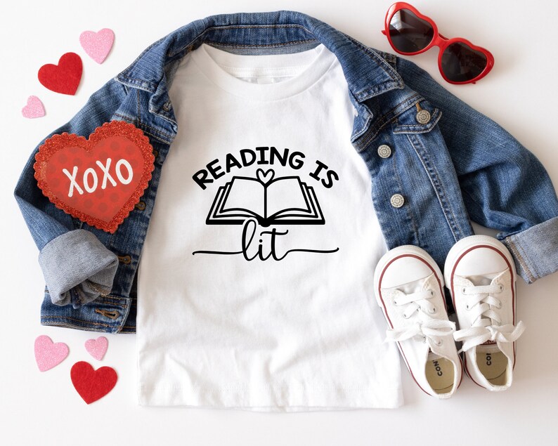 May include: White t-shirt with a black graphic that says "Reading is lit" with a book and heart design. The shirt is laid out on a white surface with a denim jacket, red heart-shaped sunglasses, and red and pink hearts.