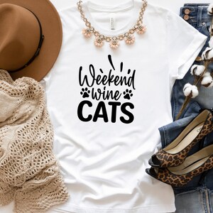 Może przedstawiać: Biała koszulka z napisem "Weekend Wine Cats" czarnymi literami. Koszulka leży płasko na białym tle z brązowym kapeluszem, dzianinowym szalikiem, parą but&oacute;w w panterkę i niebieskimi dżinsami.