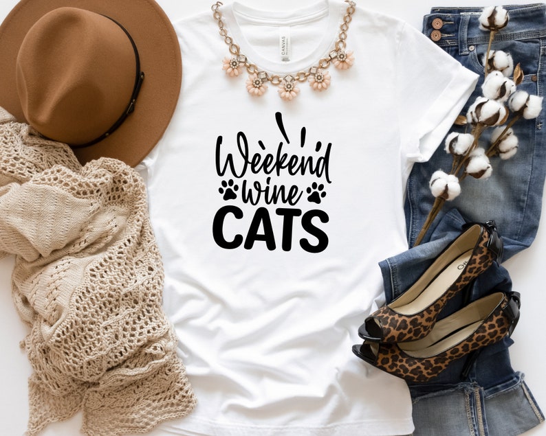 Może przedstawiać: Biała koszulka z napisem "Weekend Wine Cats" czarnymi literami. Koszulka leży płasko na białym tle z brązowym kapeluszem, dzianinowym szalikiem, parą but&oacute;w w panterkę i niebieskimi dżinsami.