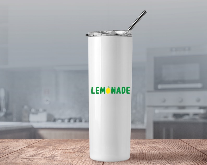 Puede incluir: Un vaso blanco de acero inoxidable con un gr&aacute;fico de "LEMONADE" verde y amarillo y una tapa plateada. El vaso est&aacute; sobre una superficie de madera.