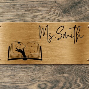 May include: A gold metal nameplate with the text "Ms. Smith" and a black and white illustration of an open book with a hand reaching for a crescent moon and stars.