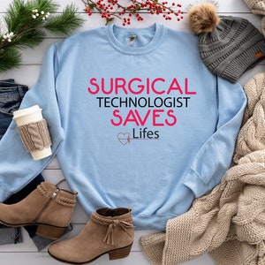 May include: Light blue sweatshirt with the text "SURGICAL TECHNOLOGIST SAVES Lifes" in pink. The text is surrounded by a pink heart with a heartbeat symbol.