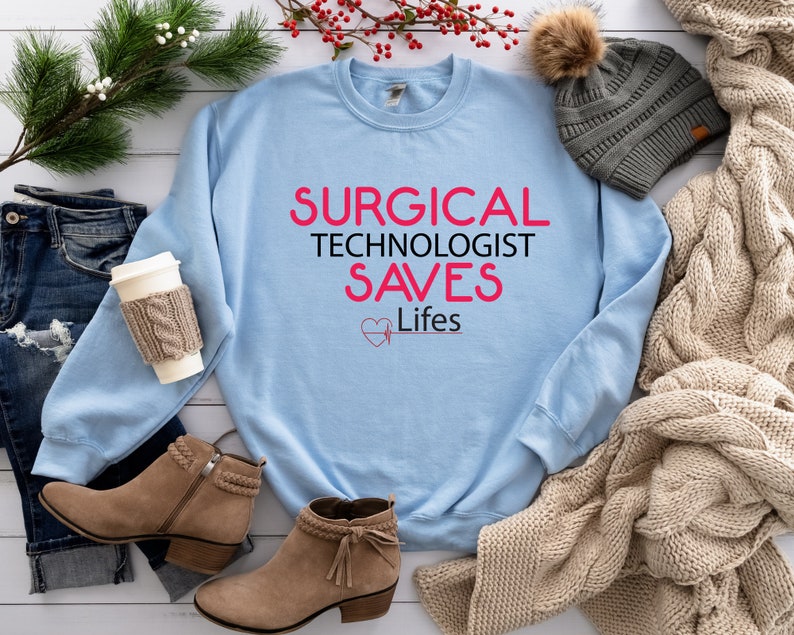 May include: Light blue sweatshirt with the text "SURGICAL TECHNOLOGIST SAVES Lifes" in pink. The text is surrounded by a pink heart with a heartbeat symbol.