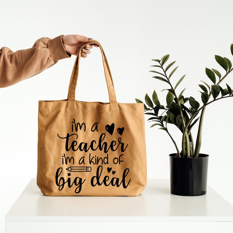 May include: A brown canvas tote bag with the text "i'm a teacher i'm a kind of big deal" in black lettering. The bag has two black hearts and a black pencil graphic.