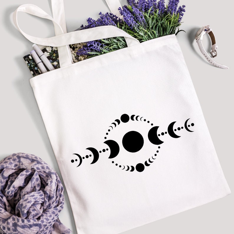 May include: White canvas tote bag featuring a black graphic of moon phases. The bag is filled with purple lavender, pens, and a floral print. A scarf and a watch are also visible.