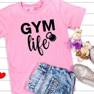 Op de afbeelding: Roze T-shirt met zwarte tekst die "GYM life" zegt en een zwarte kettlebell-afbeelding. Het shirt ligt op een wit houten oppervlak met een blauwe spijkerbroek, twee rode harten en twee knuffels.