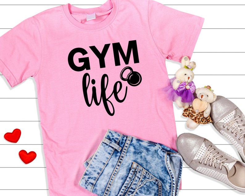 Op de afbeelding: Roze T-shirt met zwarte tekst die "GYM life" zegt en een zwarte kettlebell-afbeelding. Het shirt ligt op een wit houten oppervlak met een blauwe spijkerbroek, twee rode harten en twee knuffels.