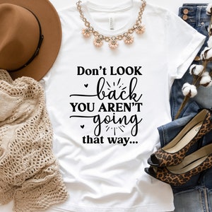 May include: White t-shirt with the text "Don't look back you aren't going that way..." printed in black. The shirt is laid flat on a surface with a brown hat, a crocheted blanket, a pair of leopard print shoes, and a pair of blue jeans.