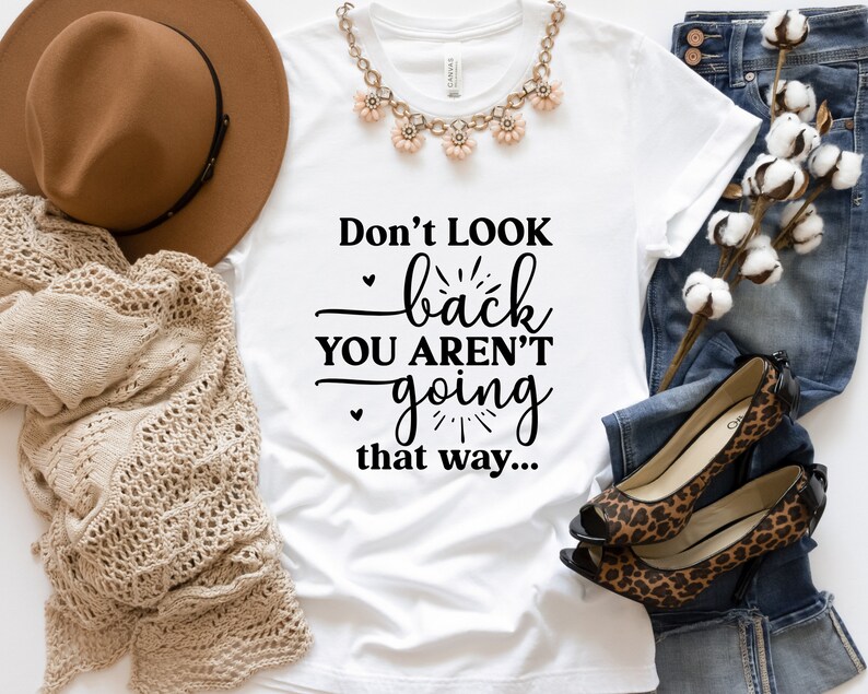 May include: White t-shirt with the text "Don't look back you aren't going that way..." printed in black. The shirt is laid flat on a surface with a brown hat, a crocheted blanket, a pair of leopard print shoes, and a pair of blue jeans.