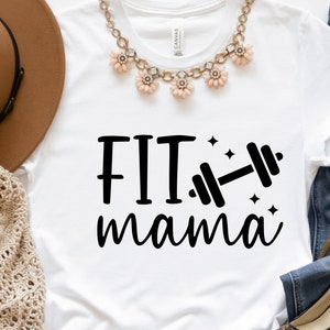 Op de afbeelding: Wit T-shirt met zwarte tekst die "FIT mama" zegt en een zwarte haltergrafiek. Het shirt ligt plat op een oppervlak met een bruine hoed, een gehaakte sjaal en een blauwe spijkerbroek.