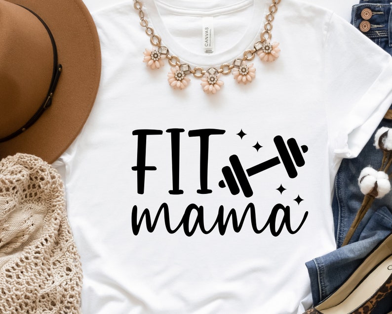 Op de afbeelding: Wit T-shirt met zwarte tekst die "FIT mama" zegt en een zwarte haltergrafiek. Het shirt ligt plat op een oppervlak met een bruine hoed, een gehaakte sjaal en een blauwe spijkerbroek.