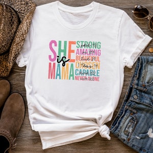 K&ouml;nnte beinhalten: Wei&szlig;es T-Shirt mit einem bunten Grafikdesign, auf dem "She is Mama" mit Bibelversen und W&ouml;rtern wie "stark", "erstaunlich", "sch&ouml;n", "auserw&auml;hlt", "f&auml;hig" und "nie allein" steht.