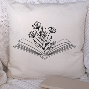 May include: A white pillow with a black line drawing of an open book with flowers growing out of it. The pillow is on a white fuzzy surface.