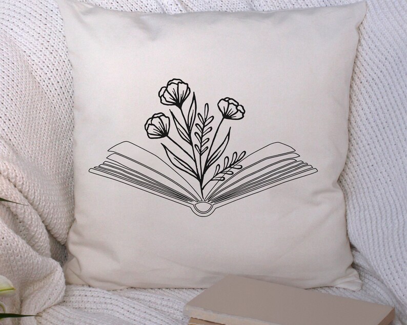 May include: A white pillow with a black line drawing of an open book with flowers growing out of it. The pillow is on a white fuzzy surface.