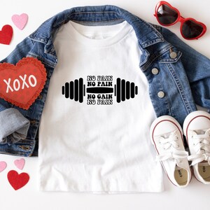Op de afbeelding: Wit T-shirt met een zwarte haltergrafiek en de tekst "No Pain No Pain No Gain No Pain". Het shirt is omringd door een spijkerjack, rode hartvormige zonnebril en rode en roze harten.