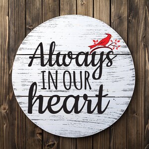 May include: White wooden round sign with distressed paint and black lettering that reads "Always in our heart". A red cardinal bird is perched on a branch with green leaves in the upper right corner of the sign.