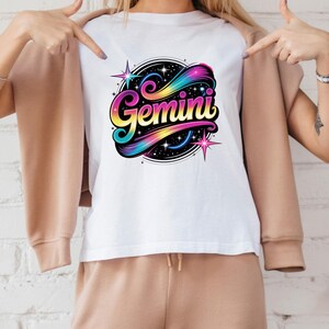 Op de afbeelding: Wit T-shirt met een kleurrijk Gemini-ontwerp. Het ontwerp bevat het woord "Gemini" in een gestileerd lettertype, omringd door sterren en een regenboogkleurige werveling tegen een zwarte achtergrond. Het is een casual kledingstuk.