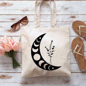 May include: Cream-colored tote bag featuring a black crescent moon design with phases and floral accents. The bag has two handles and is made of a sturdy fabric. Sunglasses and sandals are next to the bag.