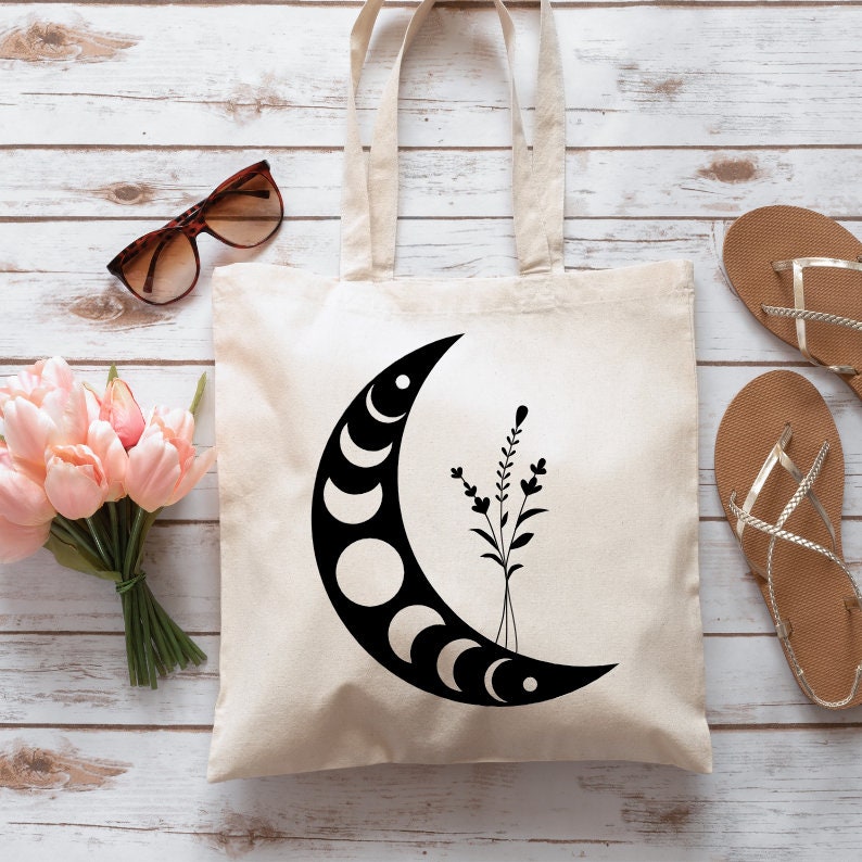 May include: Cream-colored tote bag featuring a black crescent moon design with phases and floral accents. The bag has two handles and is made of a sturdy fabric. Sunglasses and sandals are next to the bag.
