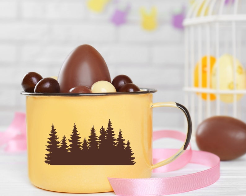 May include: A yellow ceramic mug with a forest silhouette design, filled with chocolate eggs and a larger chocolate egg on top. The mug has a handle and is sitting on a white surface with pink ribbon.