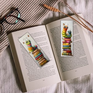 May include: Two colorful caterpillar bookmarks rest on an open book. The bookmarks feature a caterpillar design atop a stack of books. The book is open to a page with text. A pair of glasses and a sprig of wheat are also visible.