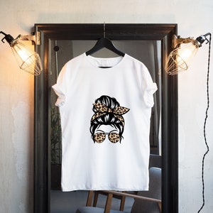 Op de afbeelding: Wit T-shirt met een afbeelding van een vrouw met een rommelige knot en een luipaardprint hoofdband met een zonnebril.