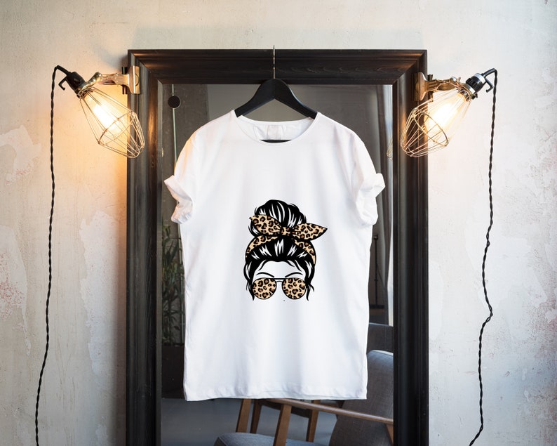 Op de afbeelding: Wit T-shirt met een afbeelding van een vrouw met een rommelige knot en een luipaardprint hoofdband met een zonnebril.