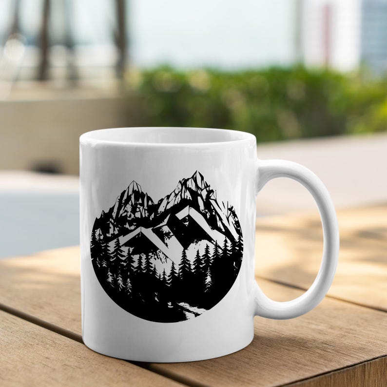 May include: White ceramic mug with a black and white illustration of a mountain range and trees in a circle design.