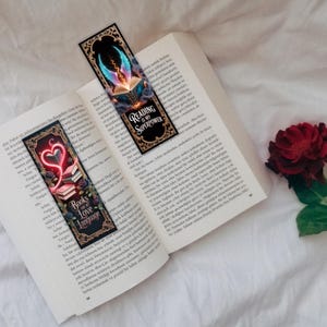 May include: Two decorative bookmarks are placed on an open book. One bookmark features a heart and books design with the text "Books are my Love Language." The other bookmark has an open book with wings and the text "Reading is my Superpower."