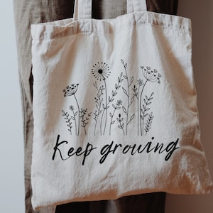 May include: A white canvas tote bag with black line drawings of wildflowers and the text "Keep growing".