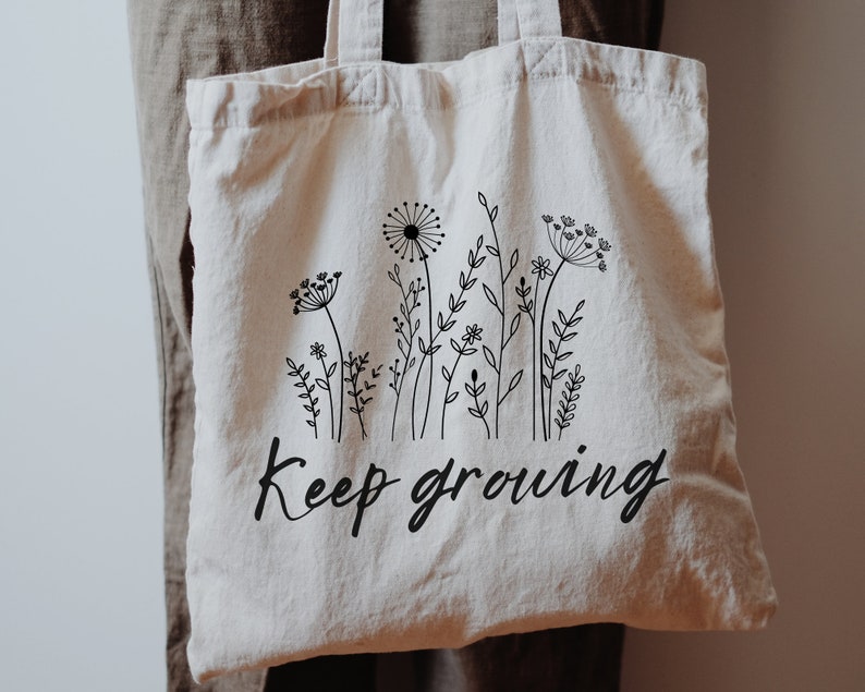 May include: A white canvas tote bag with black line drawings of wildflowers and the text "Keep growing".