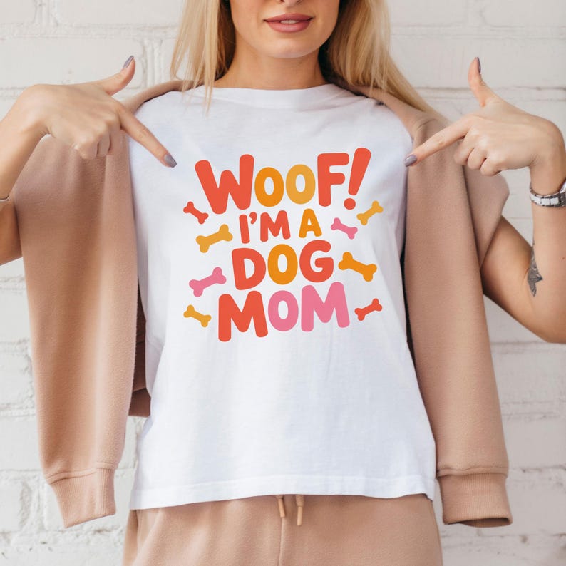 May include: White t-shirt with the text "Woof! I'm a Dog Mom" in orange and pink lettering, with bone graphics. The shirt is worn by a person with blonde hair.