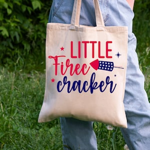 K&ouml;nnte beinhalten: Eine wei&szlig;e Canvas-Tragetasche mit dem Text "LITTLE FIRECRACKER" in Rot, Blau und Wei&szlig;. Neben dem Text befindet sich eine Grafik eines roten und blauen Feuerwerks.