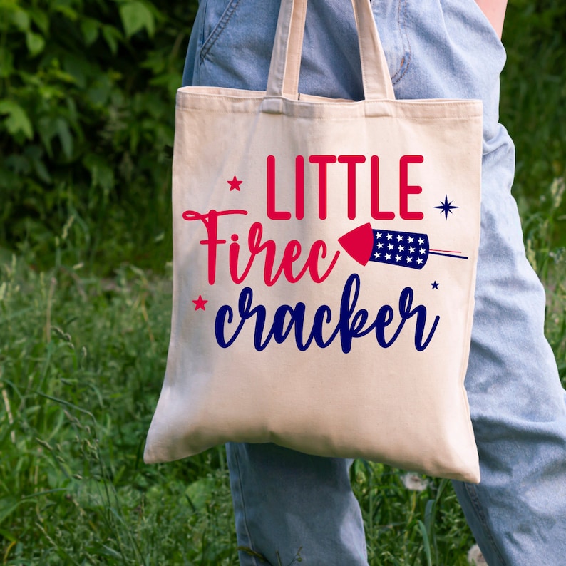 K&ouml;nnte beinhalten: Eine wei&szlig;e Canvas-Tragetasche mit dem Text "LITTLE FIRECRACKER" in Rot, Blau und Wei&szlig;. Neben dem Text befindet sich eine Grafik eines roten und blauen Feuerwerks.