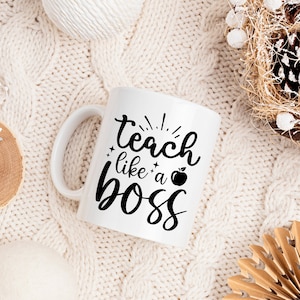 May include: White ceramic mug with black text that reads "teach like a boss" with a small apple graphic. The mug is sitting on a white knitted surface.