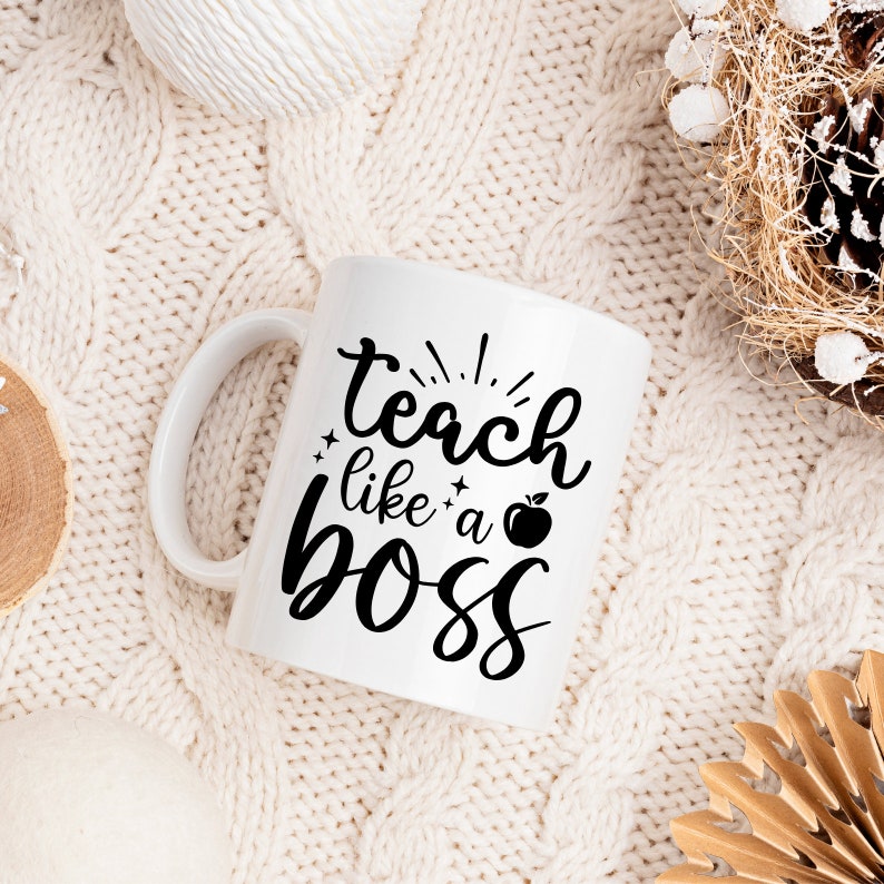 May include: White ceramic mug with black text that reads "teach like a boss" with a small apple graphic. The mug is sitting on a white knitted surface.