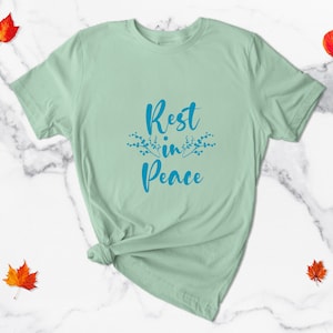 May include: A light green t-shirt with the text "Rest in Peace" in blue lettering with a floral design on either side of the text.