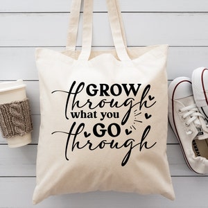May include: A white canvas tote bag with black text that reads "GROW through what you GO through" with small hearts around the text. The bag has brown straps and is sitting on a white wooden surface.