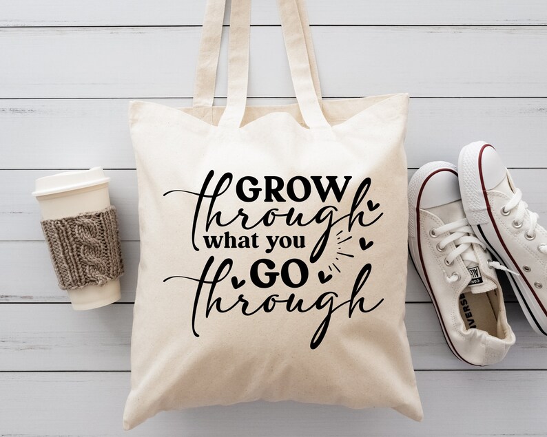 May include: A white canvas tote bag with black text that reads "GROW through what you GO through" with small hearts around the text. The bag has brown straps and is sitting on a white wooden surface.