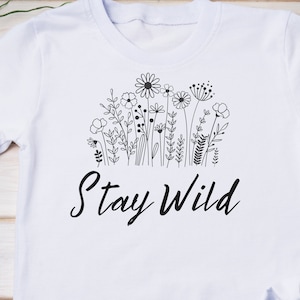 Puede incluir: Camiseta blanca con un dibujo en l&iacute;nea negra de flores silvestres y el texto "Stay Wild".