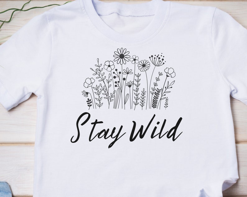Puede incluir: Camiseta blanca con un dibujo en l&iacute;nea negra de flores silvestres y el texto "Stay Wild".