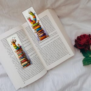 May include: Two colorful caterpillar bookmarks are placed in an open book. The bookmarks feature a stack of books with a cartoon caterpillar on top. The book is open to a page with text. A red rose is in the background.