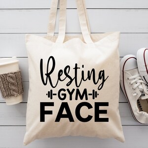 Op de afbeelding: Een witte canvas tote bag met zwarte tekst die "Resting Gym Face" zegt. De tas ligt op een wit houten oppervlak met een kopje koffie en een paar witte sneakers.