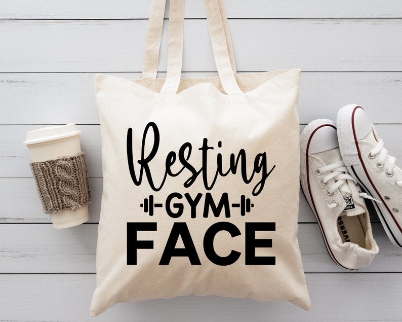 Op de afbeelding: Een witte canvas tote bag met zwarte tekst die "Resting Gym Face" zegt. De tas ligt op een wit houten oppervlak met een kopje koffie en een paar witte sneakers.
