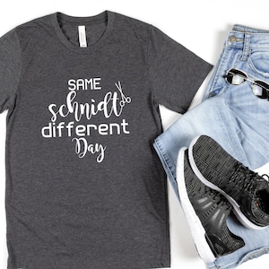 May include: A dark gray t-shirt with the text "SAME schmidt different Day" in white lettering and a pair of scissors graphic.