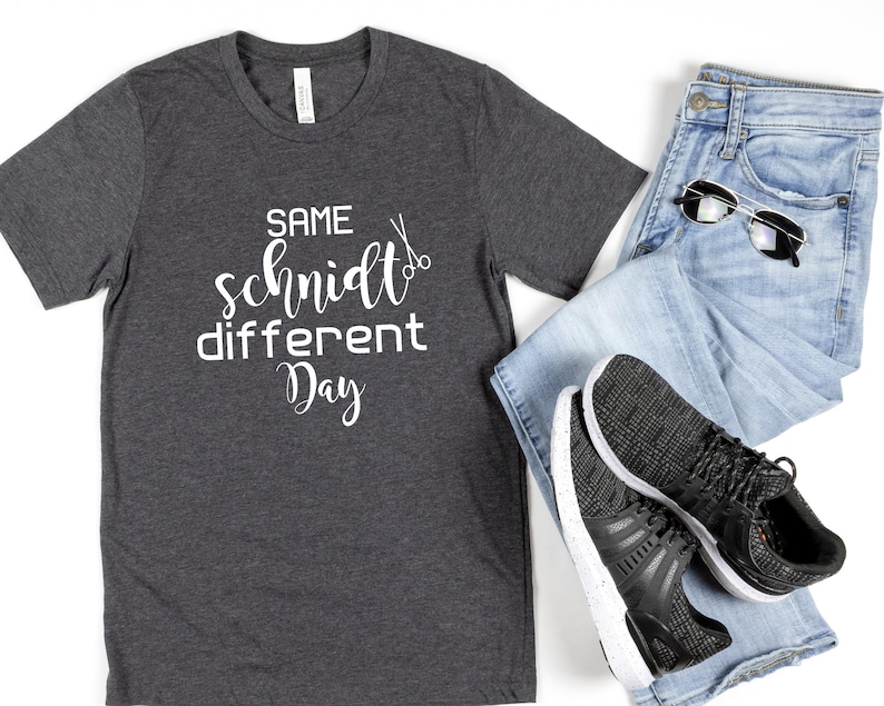 May include: A dark gray t-shirt with the text "SAME schmidt different Day" in white lettering and a pair of scissors graphic.