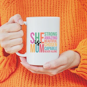 K&ouml;nnte beinhalten: Wei&szlig;e Keramiktasse mit einem bunten Design, auf dem "She is Mom" steht und W&ouml;rter wie "stark", "erstaunlich", "sch&ouml;n", "gew&auml;hlt", "f&auml;hig" und "nie allein" aufgelistet sind.
