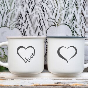May include: Two white ceramic mugs with black handles. One mug has a black heart with the word "love" written inside. The other mug has a black heart shape.