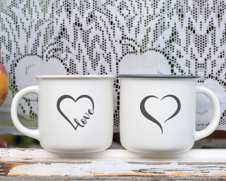 May include: Two white ceramic mugs with black handles. One mug has a black heart with the word "love" written inside. The other mug has a black heart shape.